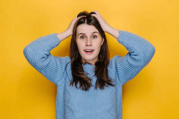 Portrait of hysterical young woman expression with her hands on head, stressed looking at camera, dressed in blue sweater, isolated on yellow studio background. People emotional lifestyle concept