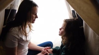 Close up of cute little smiling daughter touching foreheads with happy caring young mother, sitting together in tent. Loving two generations demonstrating support and care, enjoying tender moment.