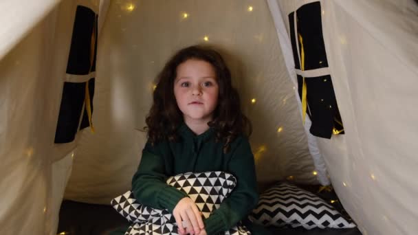 かわいいです幸せな小さな女の子ブロガー保持枕とカメラと話 小さな子供Vlogger手を振って手を見てウェブカメラ子供作るビデオ通話で座っているテントでガーランド — ストック動画