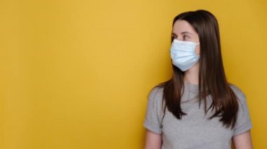 Tıbbi koruyucu yüz maskesi ve beyaz ok tutan lateks eldivenli gri tişörtlü, sarı arka planda izole edilmiş reklam alanı olan genç bir kadın. Pandemik koronavirüs konsepti