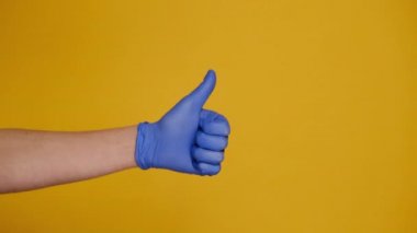 Tıbbi koruyucu mavi eldivenli erkeğe yakın çekim, baş parmak hareketi yapar, onay ya da anlaşma gösterir, sarı arka planda izole edilmiş ve reklam için fotokopi alanı ile izole edilir. El işareti. Jest gibi