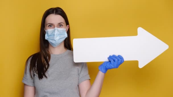 医療用マスクとラテックス手袋の若い女性は 広告のためのコピースペースと黄色の壁に隔離された灰色のTシャツに身を包んだジェスチャーのように親指を上げて矢印で脇に示しています Covid 19のコンセプト — ストック動画