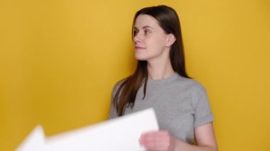 Gri tişörtlü, beyaz kağıt okla etrafı gösteren neşeli, heyecanlı genç bir kadın, reklam için fotokopi alanı olan sarı duvarlı arka plan stüdyosunda izole edilmiş. İnsanların duygu kavramı