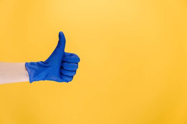 Tıbbi koruyucu mavi eldivenli erkeğe yakın çekim, baş parmak hareketi yapar, onay ya da anlaşma gösterir, sarı duvarda izole edilmiş bir şekilde reklam için yer bırakır. El işareti. Jest gibi