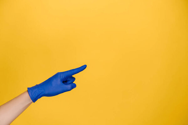 Close up male finger pointing at copy space, wears medical protective blue gloves, isolated on yellow studio background with copy space for advertisement. Advertising area, mockup. Hand gesture