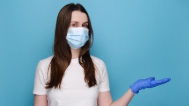Genç bir kadın kağıt beyaz ok tutuyor, kendini koronavirüsten korumak için solunum maskesi ve eldiven takıyor, tanıtım metni için boş kopya alanı gösteriyor, mavi stüdyo arka planında izole edilmiş.