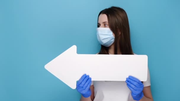 医療面のマスクと保護ラテックス手袋の紙の矢印を脇に指して若い女性の肖像画を閉じます 白いTシャツに身を包んだ 青の背景スタジオに隔離されました 流行性コロナウイルス — ストック動画