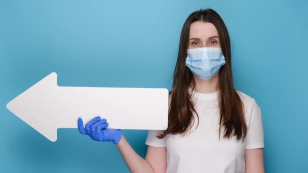 医師の顔のマスクとラテックス手袋の若い女性は ジェスチャーのような親指で脇に示す 白いTシャツに身を包んだ 広告のためのコピースペースと青のスタジオの背景に隔離された — ストック動画