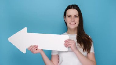 Sevimli genç bir bayan, el hareketi gibi parmakları havada, beyaz tişört giymiş, reklam için fotokopi alanı olan mavi stüdyo arka planında izole edilmiş kağıt okla bir kenara çekiliyor. İnsanların duygu kavramı