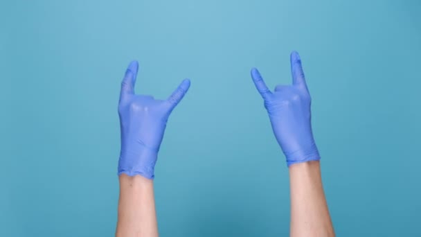 ロックンロールとパンデミックの概念 医療保護ラテックス手袋の男性の手は 広告のためのコピースペースと青のスタジオの背景に隔離された角ジェスチャーで手を振っ ボディランゲージ — ストック動画
