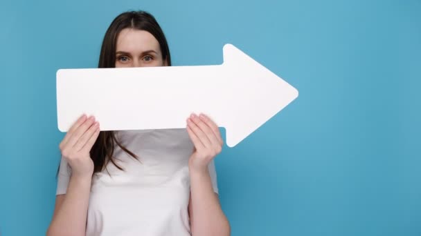 感情的な若い女性の肖像画は 広告のためのコピースペースと青の背景に隔離されたTシャツに身を包んだ白い紙の矢印とポイントの後ろに横に顔を隠します 人々の感情の概念 — ストック動画
