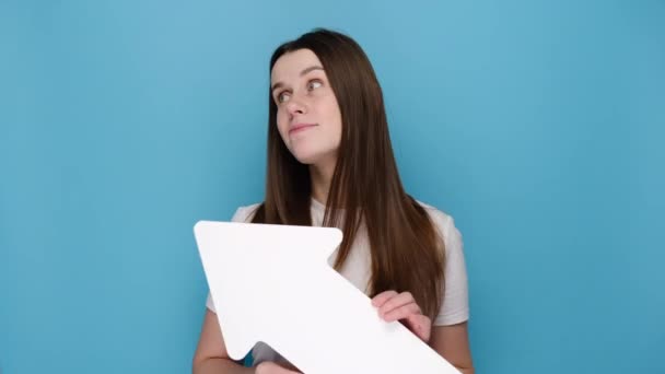 畏首畏尾的年轻女子在左上角横向握着白纸箭头 在两个选项之间做出选择 女孩耸肩 穿着T恤 与蓝色工作室背景隔离 — 图库视频影像