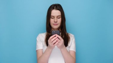 Hoş bir bayanın portresi uzak iletişimden hoşlanıyor, cep telefonu kullanıyor, hızlı ve sınırsız internet sörfü yapıyor, hoş bir gülümsemesi var, beyaz tişört giyiyor, internetten alışveriş yapıyor, mavi arka planda soyutlanıyor.