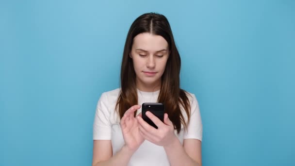 快乐的年轻女子用手机摆姿势 在现代智能手机上输入短信 手握下巴 笑得积极 在闲暇时间观看滑稽视频 被隔离在蓝色的墙上 — 图库视频影像