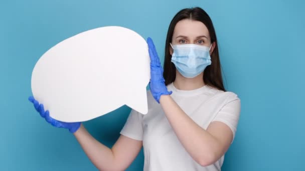 身穿医用面罩和防护手套的年轻女性 手持空白的语言泡沫 身穿白色T恤 蓝色背景隔离 并带有广告复制空间 大流行性大肠癌概念 — 图库视频影像
