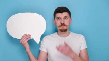 Boş konuşma balonu tutan, kollarını açıp çenesine destek olan, mavi arka plan stüdyosunda izole edilmiş beyaz tişört giyen dalgın sakallı genç adamın portresi. Duygu insanları kavramı