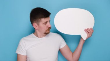 Kağıt konuşma balonu tutan genç sakallı adamın yakın plan portresi beyaz tişört giyip reklam için fotokopi çekilen mavi stüdyo arkaplanı üzerine izole edilmiş pozlar vererek iyi bir jest yapıyor. El işareti