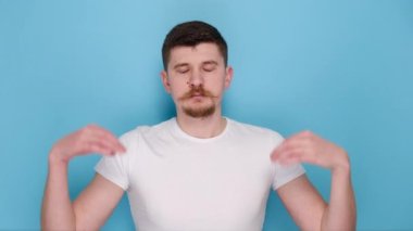 Genç sakallı adamın portresi yoga jestinde el ele tutuşuyor, dinleniyor, meditasyon yapıyor, göz kırpıyor, beyaz tişört giyiyor, mavi arka plan stüdyosunda izole edilmiş. Halk yaşam tarzı konsepti