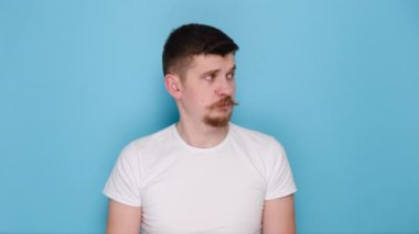Hüsrana uğramış genç, beyaz sakallı, kulakları parmakları kapalı, gözleri kapalı beyaz tişört giymiş, mavi stüdyo arka planında yalnız poz veren beyaz erkek. Duyguların insanları yaşam tarzı konsepti