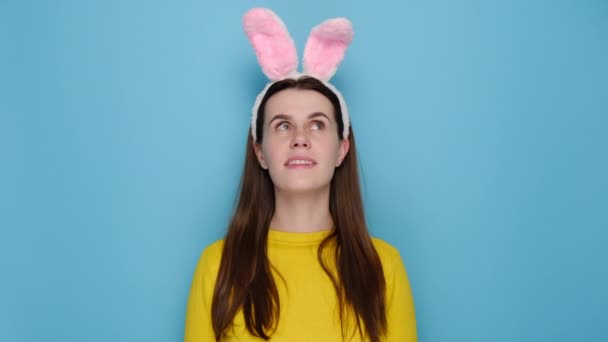 Portrait of cute smiling young woman is making wish while crossing her fingers, dressed in pink bunny fluffy ears and yellow sweater, isolated on blue studio background. Easter holiday concept