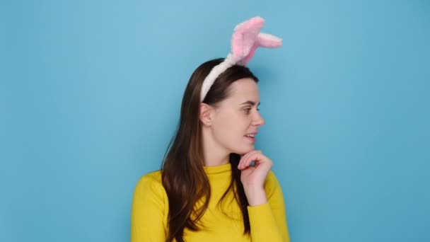 ピンクのウサギのふわふわの耳の中に魅力的な若い女性は カメラを探して 秘密のジェスチャーをしている黄色のセーターを着て 青のスタジオの背景に孤立立っている 人々の感情とイースターの概念 — ストック動画