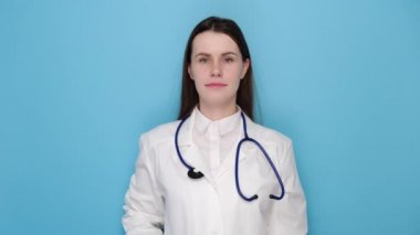 Tıbbi üniforma giyen, mutlu ve gülümseyen, mavi stüdyo arka planında izole edilmiş genç doktor portresi. Covid 19, virüs, sağlık ve tıp konsepti