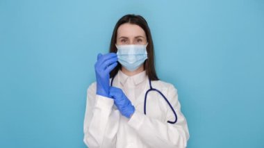 Tıbbi maskeli ve beyaz önlüklü genç bayan doktor, lastik eldiven, her şeyin yolunda olduğundan emin ol, iyi niyet göster, mavi duvarda izole edilmiş. Covid-19, sosyal uzaklık ve koronavirüs salgını konsepti