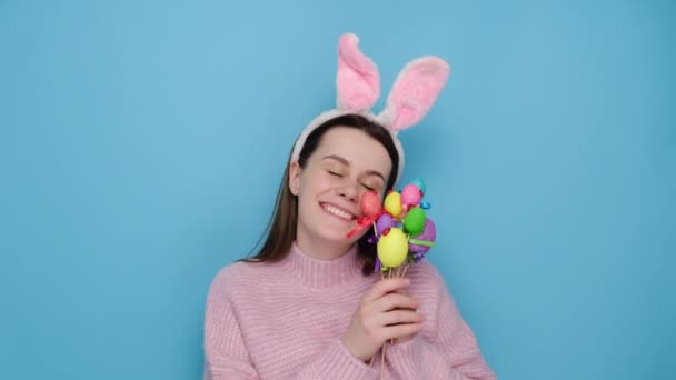 良いウサギのふわふわの耳を持つ楽観的な若い女性を見て 幸せな表情をして ピンクのセーターに身を包んだ棒に描かれた卵を保持し 青いスタジオの背景の上に立っている 春休みのコンセプト — ストック動画