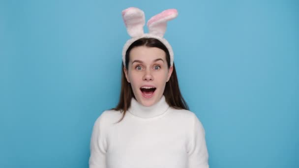 ウサギのふわふわの耳の中の驚きの女性20代の側面図は 親指を示す勝者のジェスチャーを行う頬に手を置き セーターを着て 青いスタジオの背景に隔離されました イースターの休日と感情の概念 — ストック動画