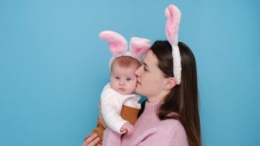 Genç, güzel bir anne, tatlı küçük kızını kucaklıyor, yanağından öpüyor ve sarılıyor, tavşan kulağı takıyor, mavi stüdyo arka planında izole edilmiş, fotokopi alanı olan. Aile ve Paskalya konsepti