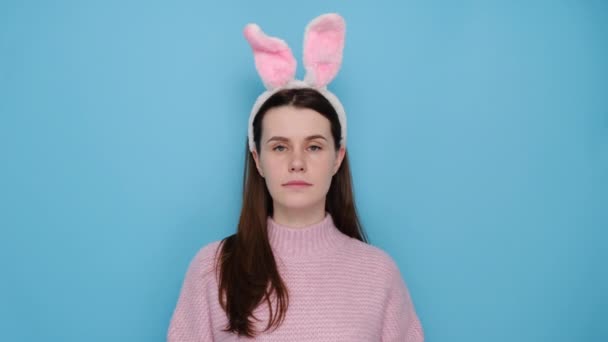 驚いたことに ウサギのふわふわの耳の中に困惑ブルネットの女性は 驚きを表現し どこでイースターを祝うために 青いスタジオの壁に隔離されたピンクのセーターを着ている不確定な感じ 春休みのコンセプト — ストック動画