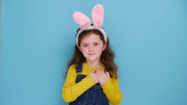 Pembe tüylü kulaklı, gülümseyen kreş çocuğu portresi mavi stüdyo arka planında izole edilmiş, katlanmış avuç içlerini kalbe koydu. Paskalyayı kavra ve içten bir insan olmanın işareti, minnettar hisset.