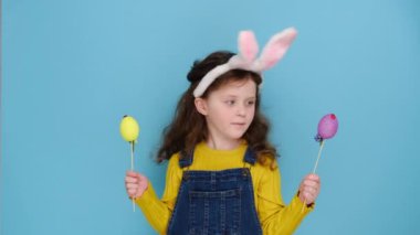 Pembe tüylü tavşan kulaklı, elinde iki renkli yumurta tutan, dini bayramı simgeleyen, mavi stüdyo arka planında izole edilmiş, anaokulu öğrencisi bir kız çocuğu. Çocukluk yaşam tarzı ve Mutlu Paskalyalar konsepti