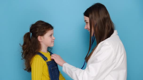 聴診器を使用している女性医師小児科医は 幸せな健康的なかわいい就学前の子供の女の子の心を聞いて 青い壁に隔離された医療訪問で子供の患者に高い5を与えます 子どもの健康コンセプト — ストック動画