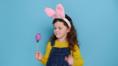 Pozitif küçük kız pembe tavşan kulağı takıyor ve kazak giyiyor, küçük renkli yumurtalar büyüyor, tatil süslemeleri yapıyor, mavi stüdyo arka planında modeller yapıyor. Şu süslü Paskalya yumurtalarına bak.!