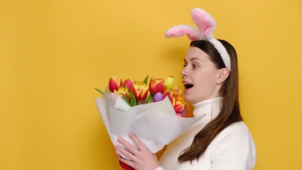 有名なピンクのウサギのふわふわの耳に若い女性の窒息春の花の花束をキャッチカラフルな小さな卵が飾られ 興奮ああ 黄色のスタジオの背景に隔離された私の神すごいと言う イースターのコンセプト — ストック動画