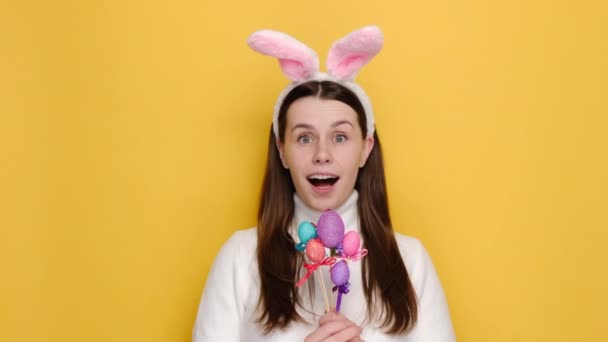 ピンクのウサギのふわふわの耳に笑顔の若い女の子20代の肖像画は イースターエッグを取ることを示唆し 手に象徴的なものを持って 黄色のスタジオの背景に白いジャンパー モデルを身に着けています 休日のコンセプト — ストック動画