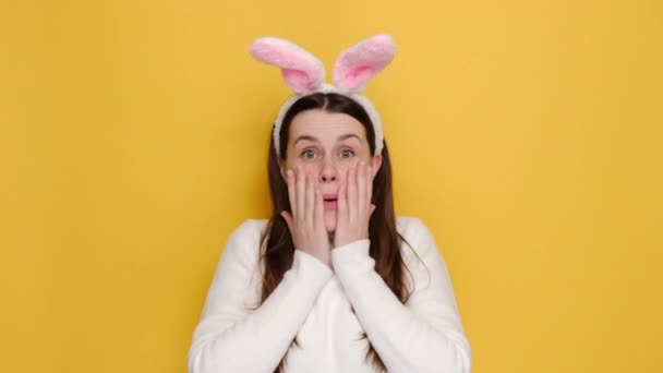 ピンクのウサギのふわふわの耳に驚きショックを受けた若い女性20代の頬の頭の上にすごい手を入れて 黄色のスタジオの背景に隔離された白いジャンパーを着ていると言う イースター休暇と人々の感情の概念 — ストック動画