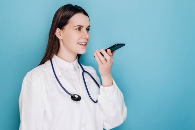 Beyaz tıbbi üniforma giyen ve steteskop tutan gülümseyen kadın doktor akıllı telefondan sanal dijital ses asistanını etkinleştir, hastaya internetten danış, mavi arka planda izole ol.