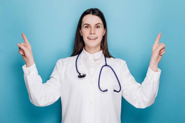 Dikkatli hemşire, palto ve steteskop giyen doktor, üst tarafı işaret eden, mavi duvarda izole edilmiş. Hekim gülümseyerek müthiş bir duyuru yaptı. Covid 19, sağlık çalışanları, salgın konsepti.