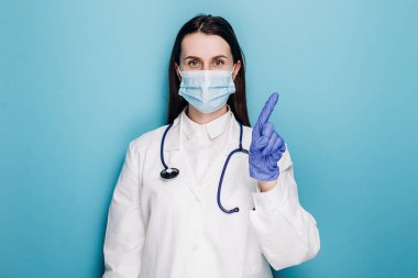 Koruyucu maskeli ve eldivenli ciddi bir profesyonel kadın doktor insanları uyarıyor. Yasaklama kısıtlamasında parmağını sallıyor, mavi duvarda izole edilmiş. Tıp çalışanları, pandemik koronavirüs konsepti