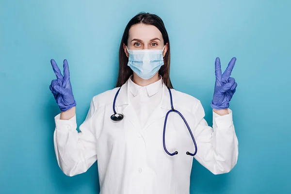 Optimistic cute woman doctor ask to stay safe, wear medical mask and latex gloves, show peace sign, hope we defeat virus pandemia, isolated on blue wall. Covid 19, healthcare and doctors concept