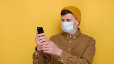 Milenyum Kafkasyalı tıbbi maskeli bir adam akıllı telefondan video görüşmesi yapıyor, genç bir erkek web kameralı dijital konuşma yapıyor ya da cep telefonuyla sanal konferans yapıyor, sarı duvarda izole edilmiş.