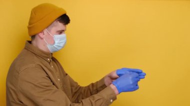 Tıbbi maske takan genç adam tıbbi eldivenlerini çıkarıp çöpe atıyor, gömlek ve bere giyiyor, sarı stüdyo arka planında izole edilmiş ve fotokopi alanı olan bir şapka giyiyor. Virüs salgınının sonu kavramı