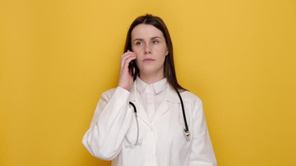 怒り狂った若い女性医師や看護師が携帯電話で話していると 悲鳴を上げる普及手を身に着けているものを尋ねます 白いコートと聴診器で身に着けています 黄色のスタジオの背景に隔離 — ストック動画