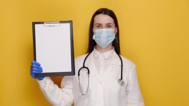 Orvosnő Orvosi Maszkban Kesztyű Papírra Mutatva Kabátot Sztetoszkópot Visel Sárga — Stock videók