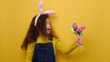 Pozitif küçük kız, pembe tavşan kulağı takıyor, küçük renkli yumurtalar büyüyor, baş parmağını gösteriyor, jest gibi, onayını gösteriyor, sarı duvarın üzerinde izole edilmiş. Şu süslü Paskalya yumurtalarına bak.!