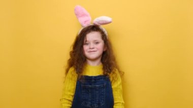 Kamerayı gösteren tüylü tüylü kulaklı, birinci sınıf jestler yapan, süveter giyen, sarı arka planda izole edilmiş okul öncesi küçük kız. Paskalya bayramı ve duygu konsepti