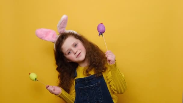 有趣的学龄前小女孩积极地看待相机 拿着两个复活节彩蛋盖住眼睛 是兔子 黄色工作室背景的模特 复活节和季节性假日的快乐概念 — 图库视频影像