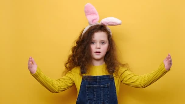 ピンクのウサギのふわふわの耳の手で大小のサインを示すジェスチャーで魅力的な子供の女の子 メジャーシンボル カメラを見てショックを受け 黄色の壁に隔離された イースターと測定の概念 — ストック動画